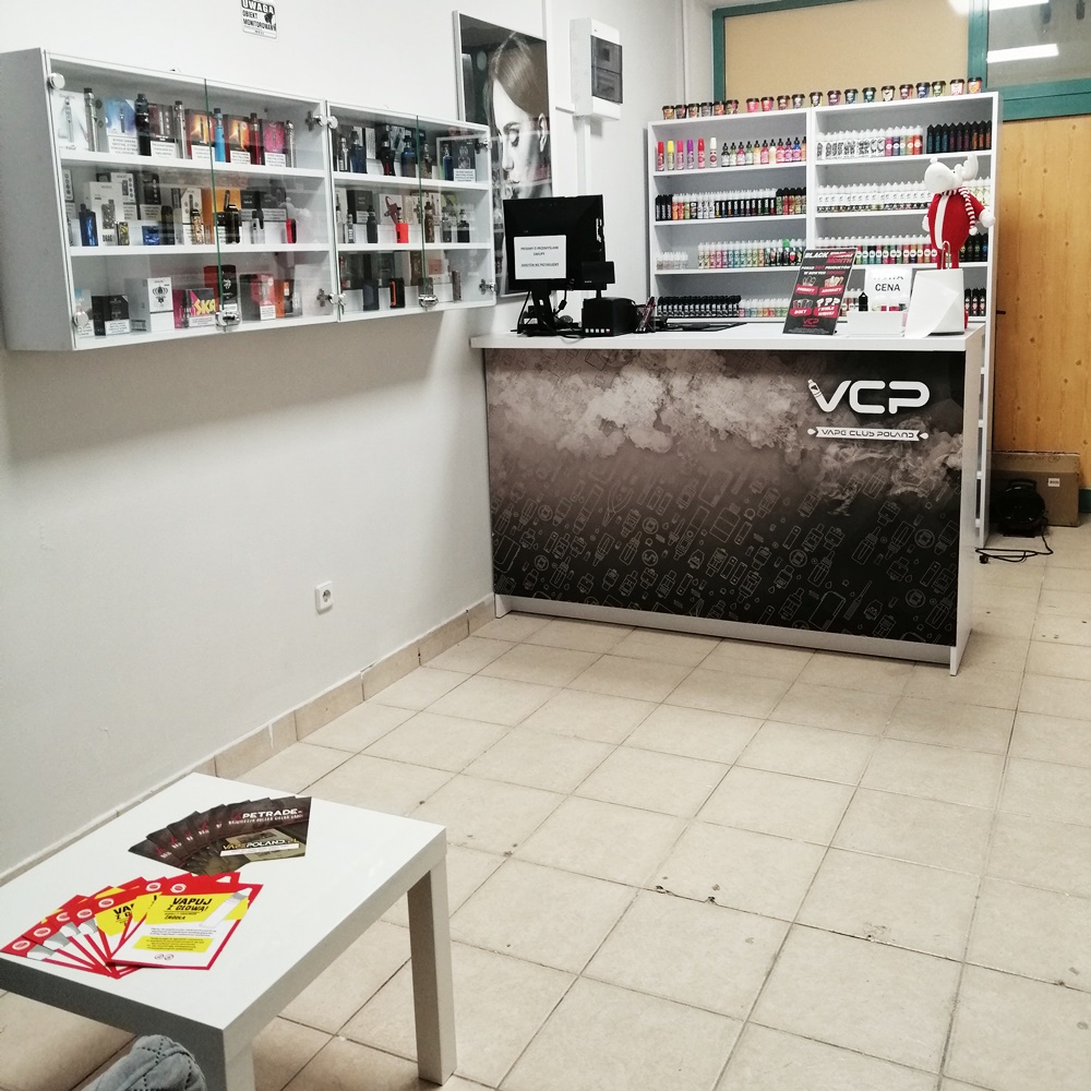 Wnętrze stacjonarnego sklepu z e-papierosami, charakteryzujące się sterylną bielą, jasnym oświetleniem LED i sprzętem vape elegancko wyłożonym za szklanymi witrynami, przypominające aptekę lub salon Apple Store.