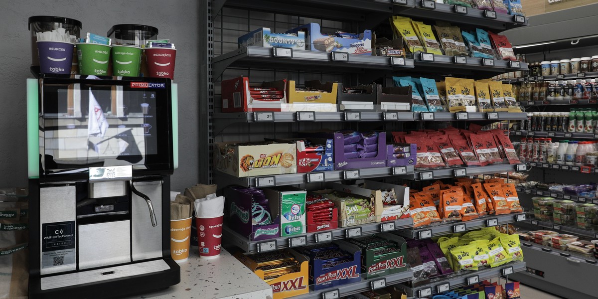 Zbliżenie na bardzo zatłoczoną ladę kasową w sklepie Żabka, wypełnioną drobnymi produktami impulsowymi, podkreślające walkę o każdy centymetr przestrzeni.